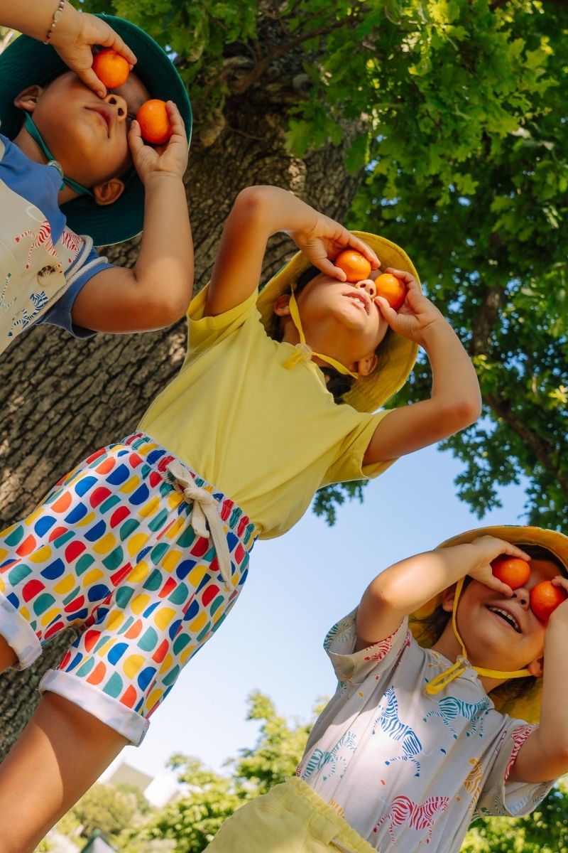Three children in colorful hats holding fruits under a tree on a sunny day