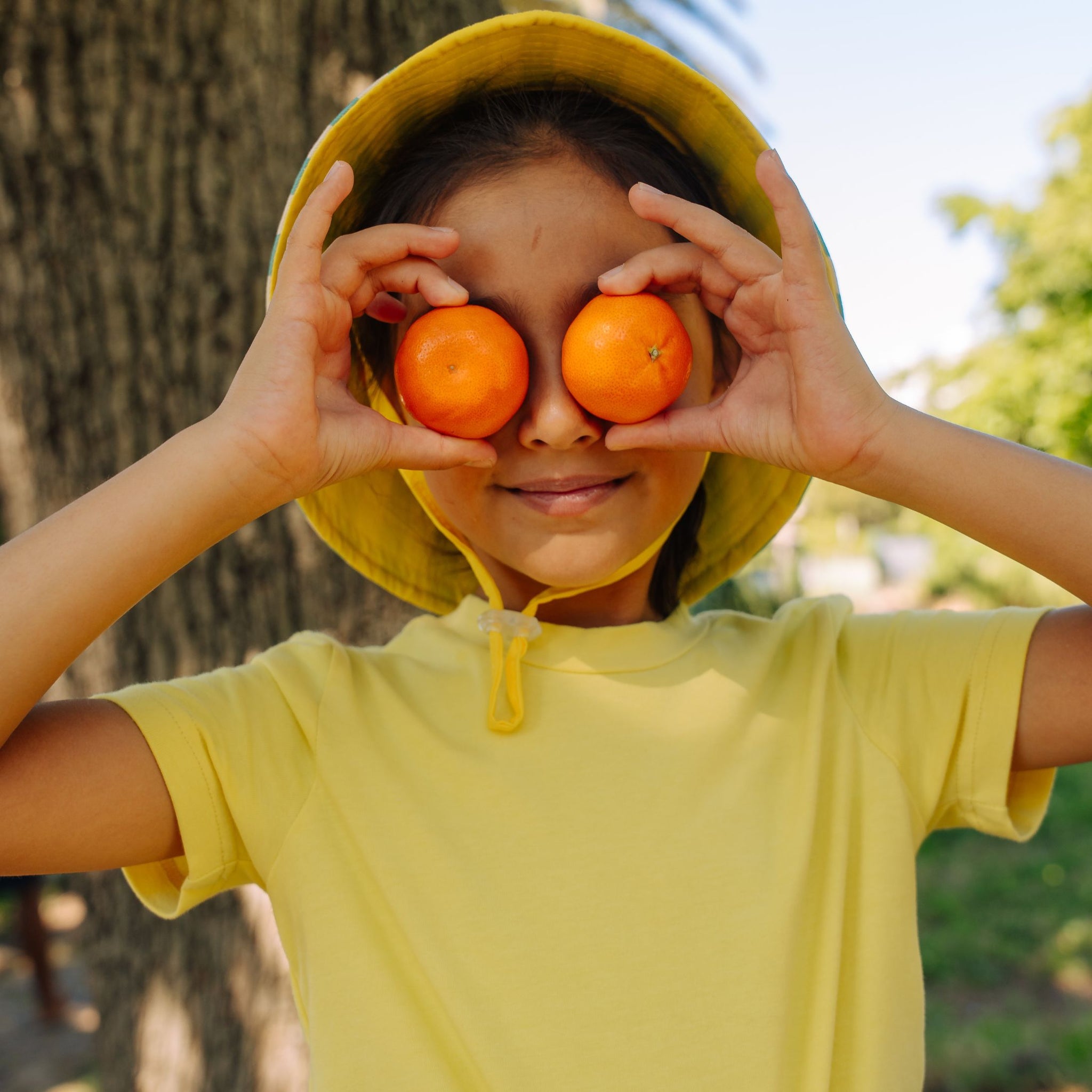 Girl smiling and holding clementines in front of her eyes wearing Sunkin ts-hirt in yellow