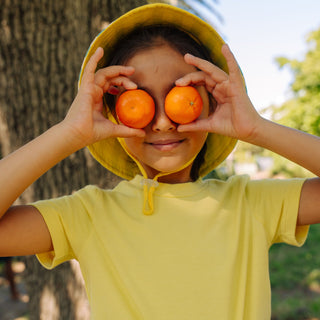 Girl smiling and holding clementines in front of her eyes wearing Sunkin ts-hirt in yellow