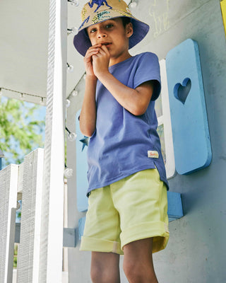 Child wearing a purple tee shirt and yellow shorts standing outdoors near a white structure with blue heart-shaped cutout.