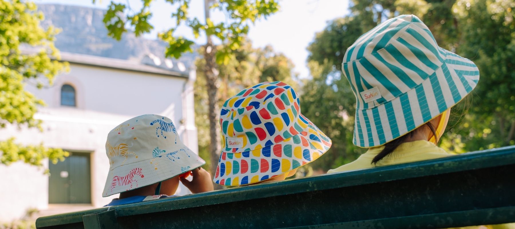 Three children wearing colorful SunKin bucket hats sitting on a bench outdoors.