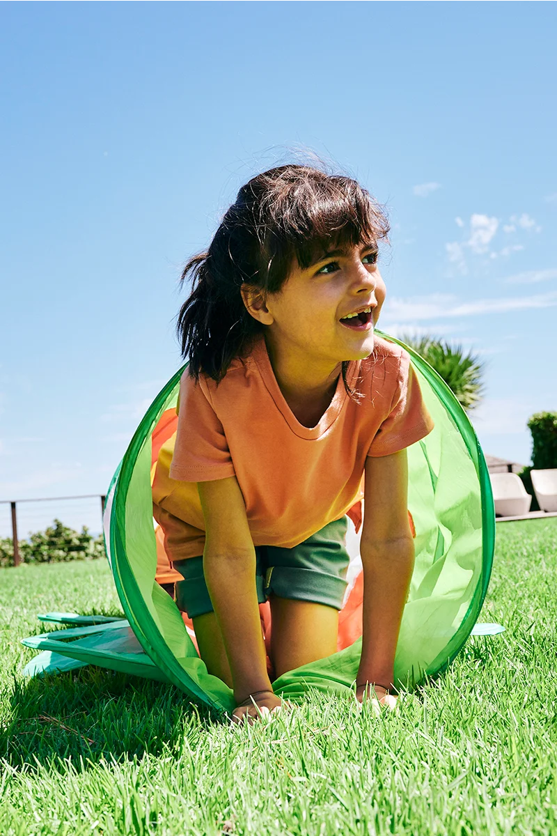Child wearing coral SunKin shirt crawling through a green tunnel on a sunny lawn in UPF50+ clothing - MOBILE version