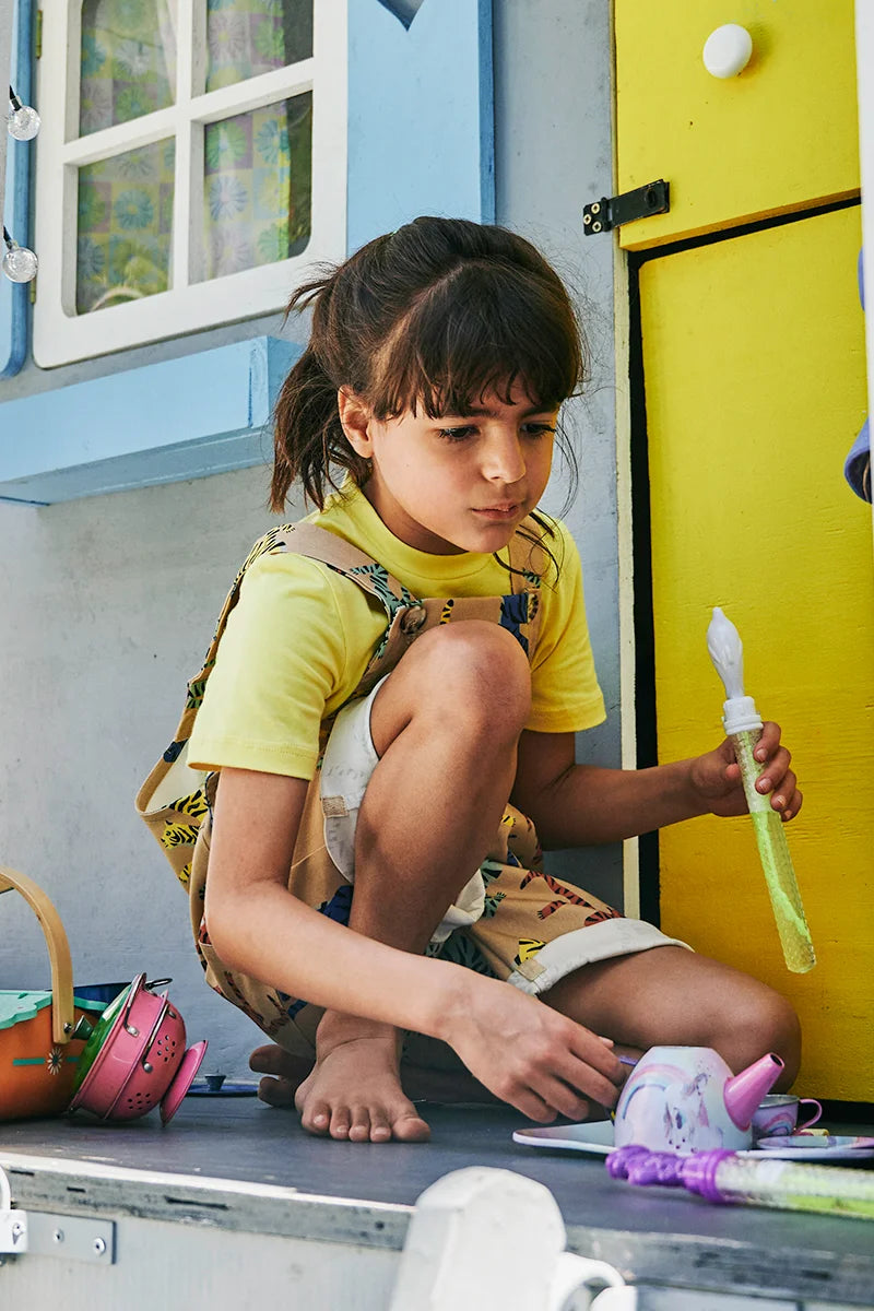 Child in yellow SunKin shirt and printed dungarees playing with bubble toys outside a white playhouse. MOBILE version