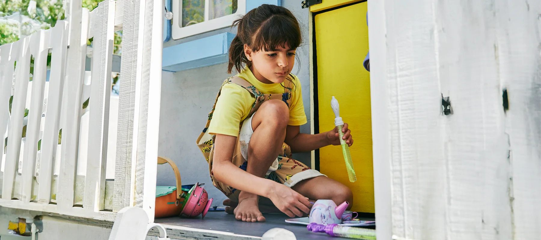 Child in yellow SunKin shirt and printed dungarees playing with bubble toys outside a white playhouse
